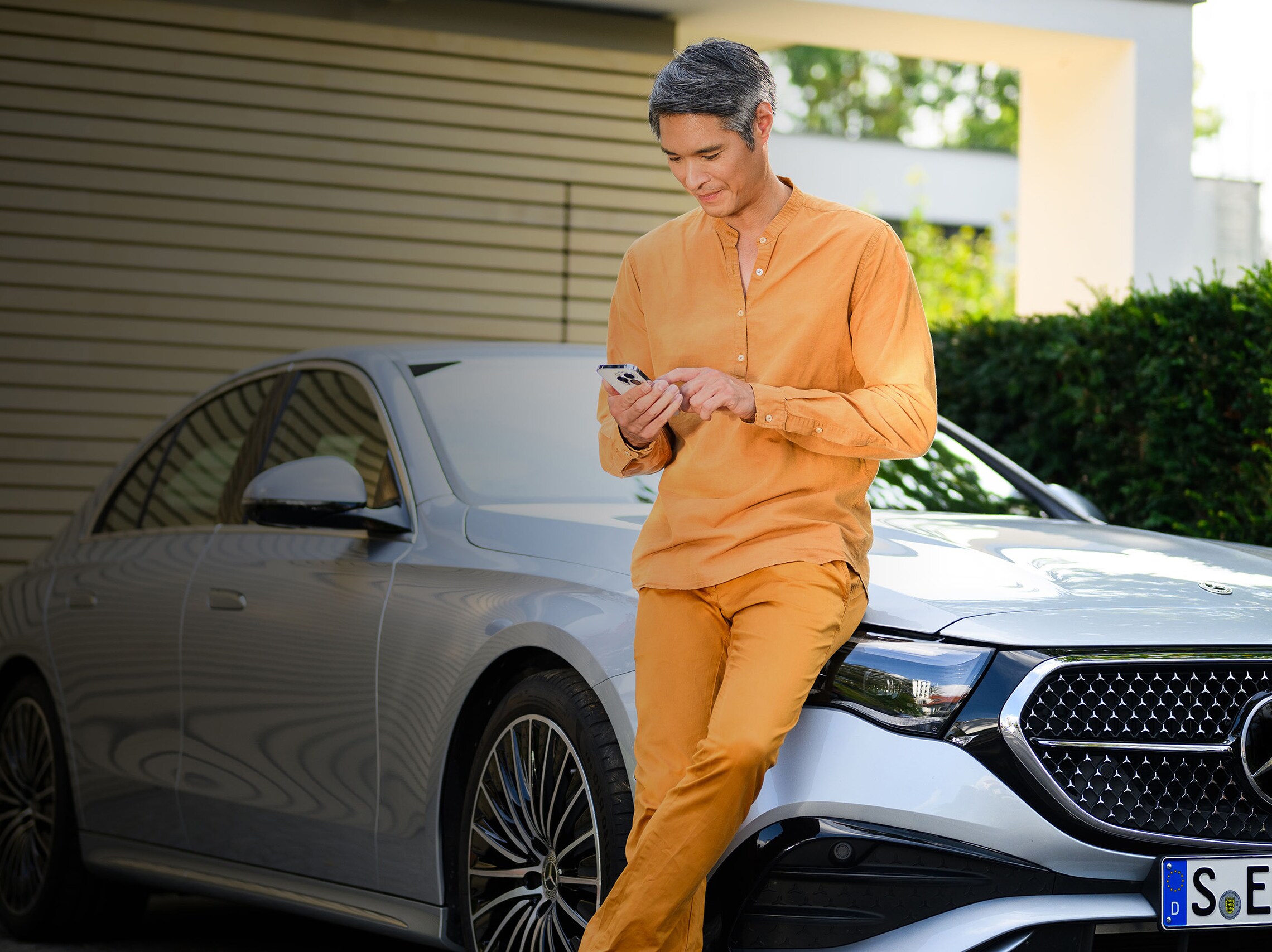 A person leaning against a Mercedes-Benz vehicle, using a mobile device. 