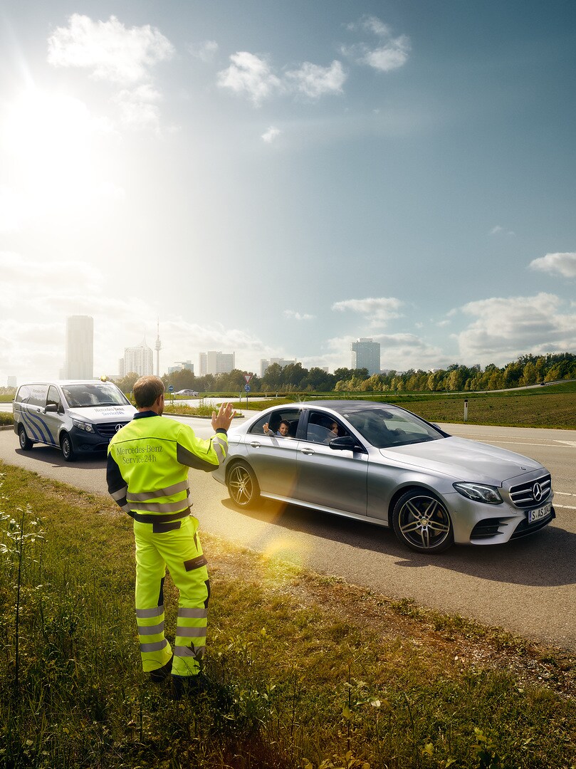 A Mercedes-Benz service technician waving from the roadside to a child sitting in an approaching Mercedes.