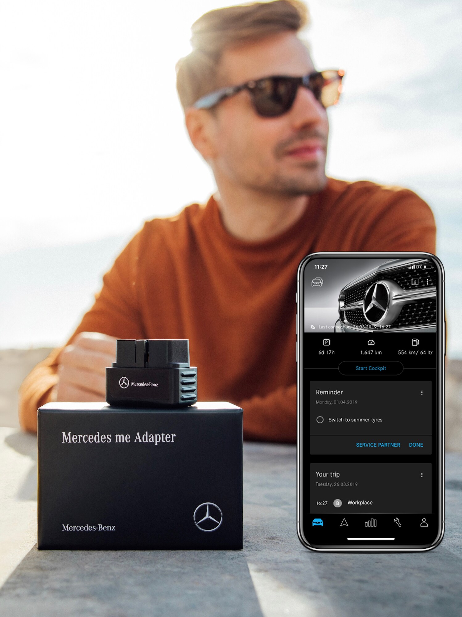 A man wearing sunglasses sitting at a table with a Mercedes-Benz Adapter and a smartphone with the Mercedes-Benz Adapter app open in the foreground.