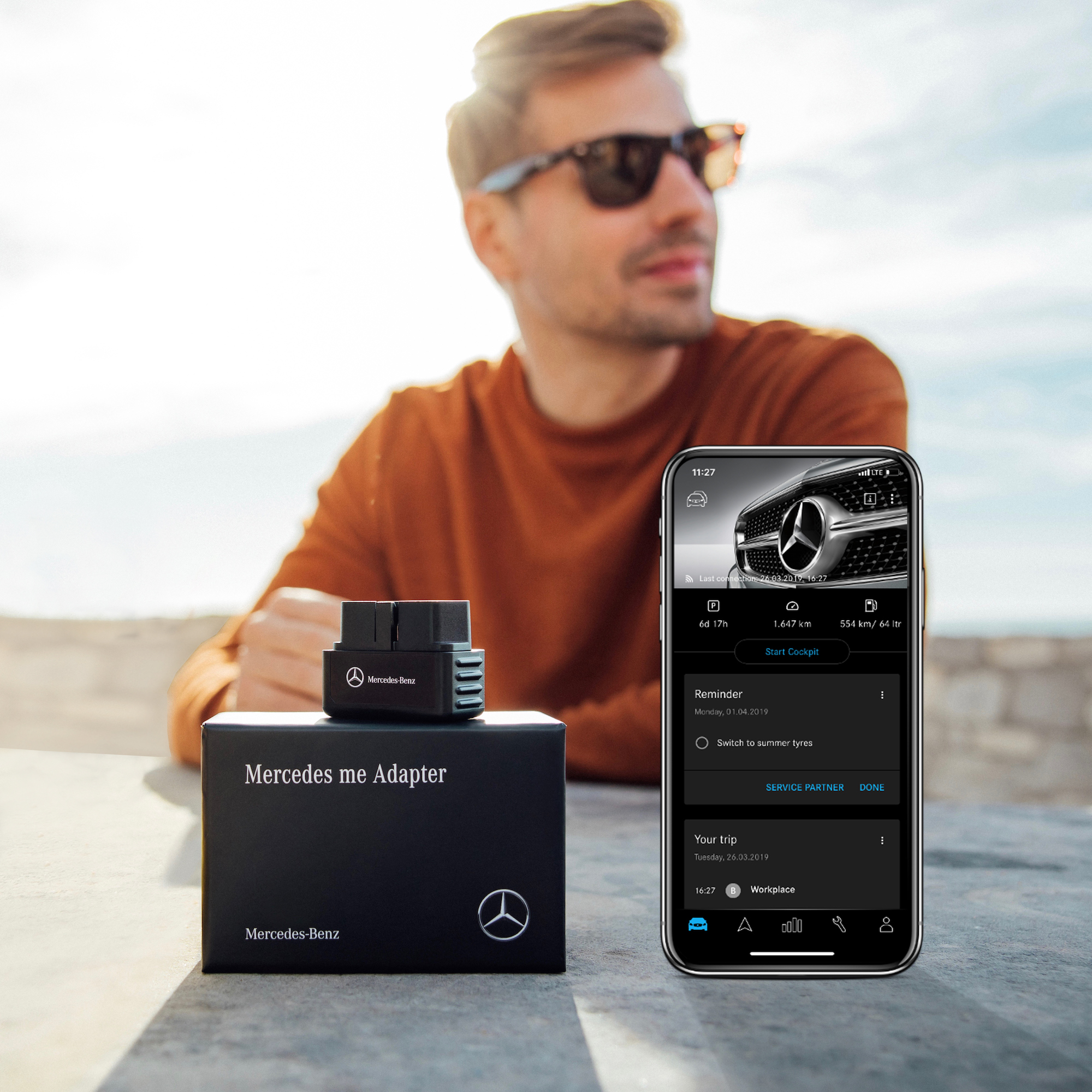 Accident detection with the Mercedes-Benz Adapter. | Mercedes-Benz A man wearing sunglasses sitting at a table with a Mercedes-Benz Adapter and a smartphone with the Mercedes-Benz Adapter app open in the foreground.