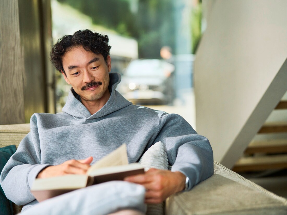 Customer sitting indoors reading a book. Visible through a window behind him, a car is being serviced by a specialist.