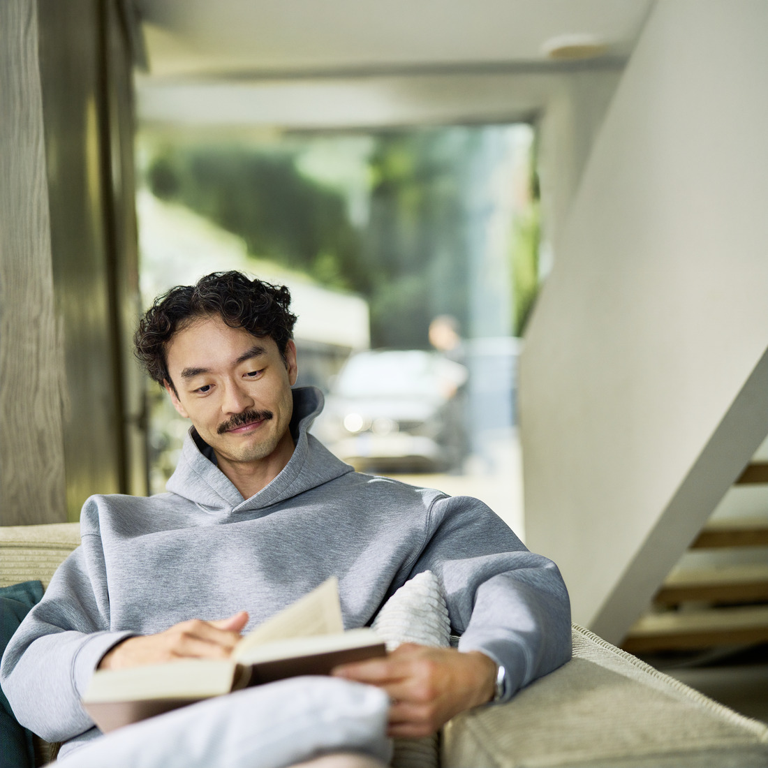 Relaxed customer reciving Mobile Service at their home Customer sitting indoors reading a book. Visible through a window behind him, a car is being serviced by a specialist.